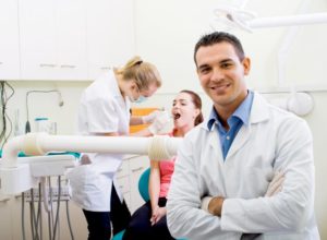 A dentist smiling while his colleague examines a patient.